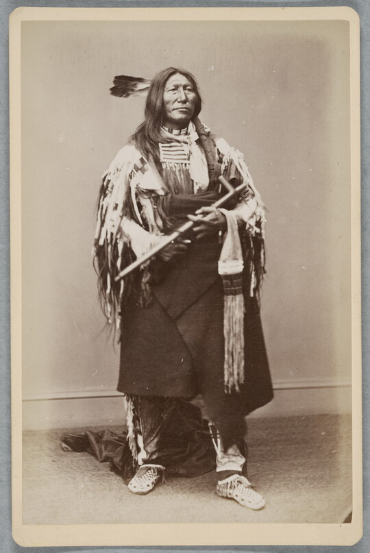 A sepia-toned full-body portrait of an Indigenous man in cultural dress holding a pipe.