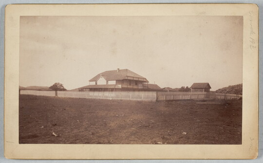 A sepia-toned photograph of a two-story house behind a picket fence on an open field.