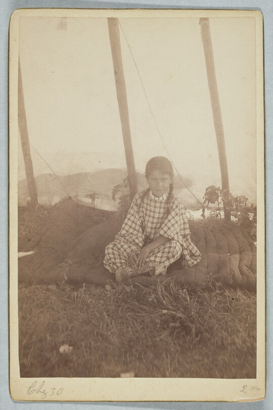 A sepia-toned photograph of a young girl wearing a gingham dress and sitting cross-legged on a blanket.
