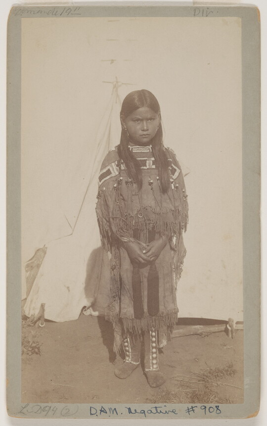 A sepia-toned photograph of a young Native American girl in traditional clothing standing outside.