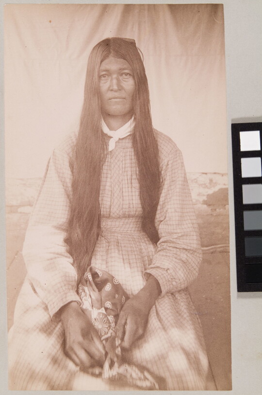 A sepia-toned portrait of a seated Indigenous woman with long dark hair wearing a long-sleeved gingham dress.