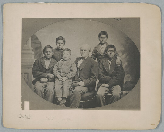 A black-and-white studio portrait photograph of a seated White man surrounded by five Indigenous boys of various ages.