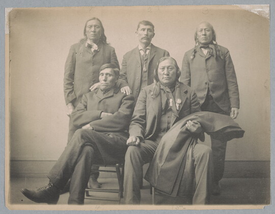 A sepia-toned studio portrait photograph of five Indigenous men in dark suits, three standing and two seated.