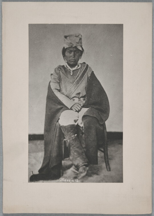 A black-and-white portrait photograph of a seated Native American man wearing a head covering and with a blanket wrapped around is shoulders.