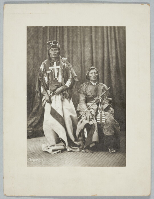 A black-and-white portrait photograph of two Indigenous adults, one standing and one sitting, wearing traditional clothing, one holding a pipe.