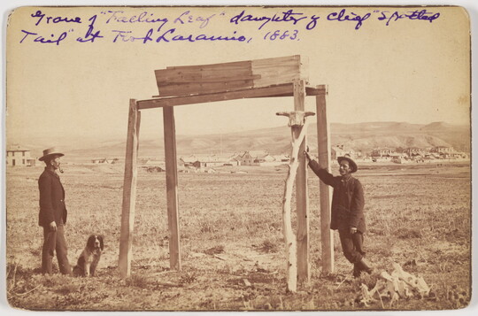 A sepia-toned photograph of two men and a dog next to a simple wood structure on the outskirts of a town.