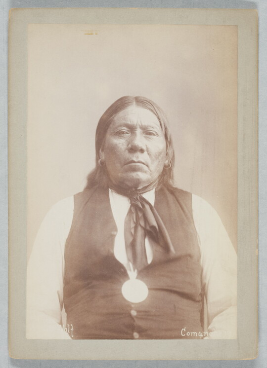 A black-and-white portrait photograph of an Indigenous person with long hair and earrings wearing a white shirt under a buttoned vest.