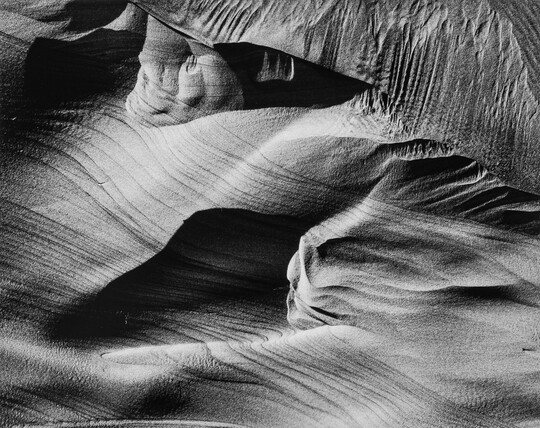 An abstract black-and-white photograph of sand dunes with horizontal and vertical striations that make it seem to be in motion.