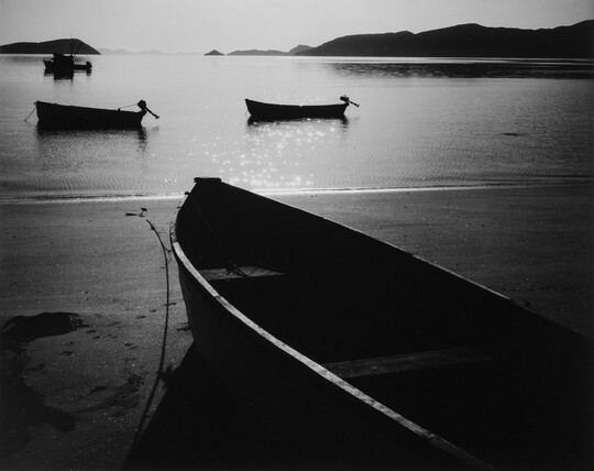 A black-and-white photograph of a rowboat on a sandy beach and three more out on still water with hills in the distance.
