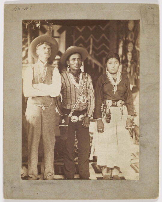 A sepia-toned photograph of three figures [L-R]: a man in western dress, a man wearing a concho belt over western clothing, and a woman in a skirt and blouse with a concho belt.