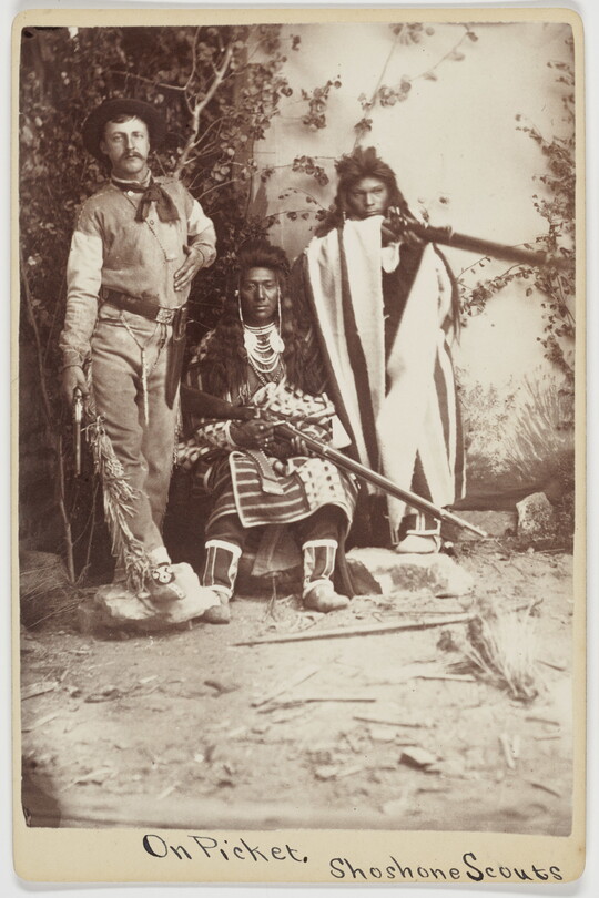 A sepia-toned photograph of three people, one dressed in western attire with a pistol at his side, and two in Indigenous people in cultural dress holding rifles.