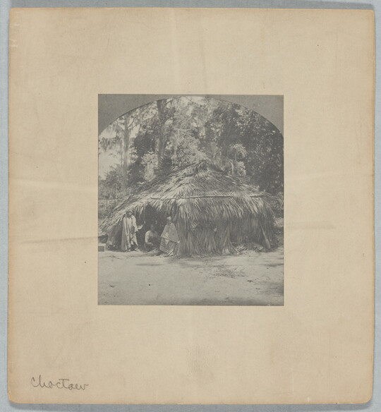 A black-and-white photograph of a wigwam with three figures in the doorway.