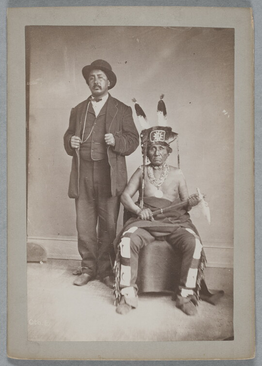 A black-and-white photograph of two Indigenous men: the one standing is wearing a bowler hat and a three-piece suit; the one seated wears a feathered hat, many necklaces, fringed pants, and holds a tomahawk in his lap.