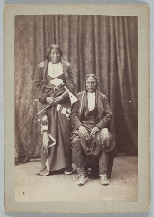 A sepia-toned portrait photograph of two Indigenous people: one seated with a hat on their knee, and the other standing holding a tomahawk.