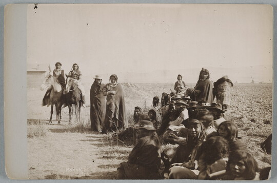 A sepia-toned photograph of a group of mostly seated Indigenous figures and two on horseback. 