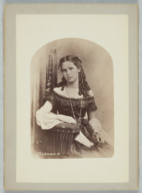 A sepia-toned portrait of a light-skinned teenage girl, seated in a chair, wearing a formal, off-the-shoulder dress with her hair styled in ringlets that fall over her shoulder.