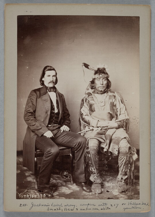 A sepia-toned portrait photograph of two seated men: a White man wearing a long suit jacket over a vest, and the other an Indigenous man in traditional dress holding a pipe.