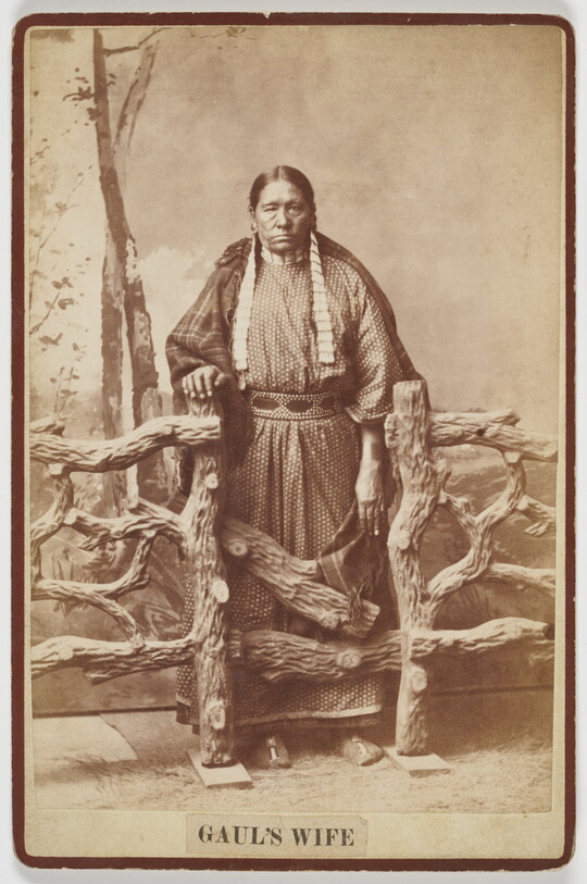 A sepia-toned studio photograph of an older Indigenous woman wearing a long polka-dotted dress, blanket around her shoulders, standing behind a wood fence.