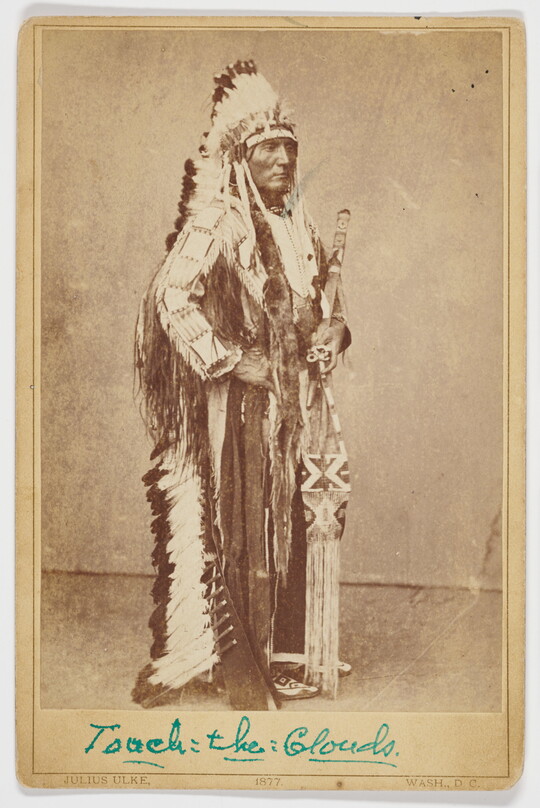 A sepia-toned portrait photograph of an Indigenous chief standing in traditional regalia, including a full-feathered headdress.