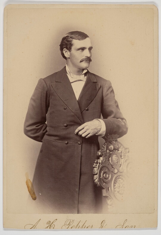 A sepia-toned portrait of a White man wearing a long frock coat standing posed with his arm on the back of a chair.