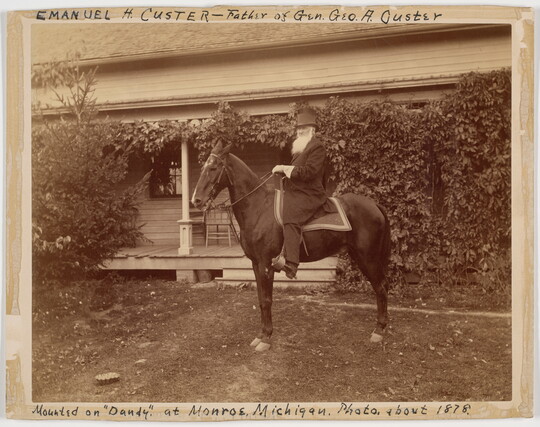 A sepia-toned photograph of a bearded White man seated on a horse in front of a house covered in vines.