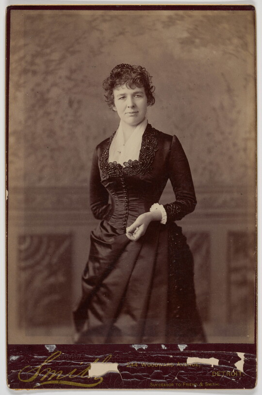 A black-and-white studio portrait photograph of a White woman in a formal, fitted dress standing in front of a backdrop.