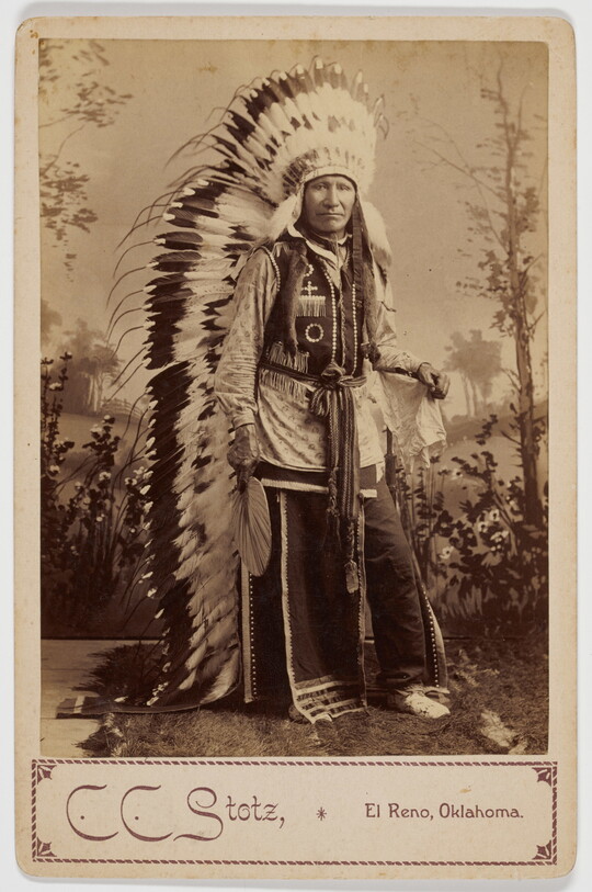 A sepia-toned studio portrait photograph of an Indigenous man in full traditional regalia, including a long, feathered headdress.
