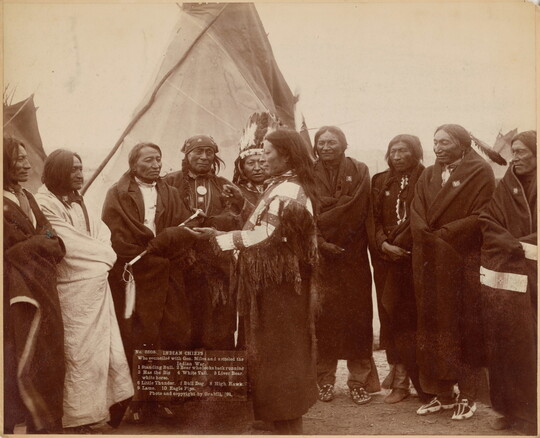 A sepia-toned photograph of Indigenous men wrapped in blankets meeting in front of a tipi.