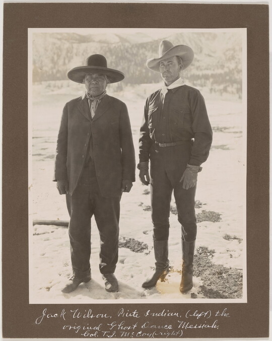 A black-and-white photograph of an Indigenous man in a suit and wide-brimmed hat standing next to a White man in dark clothes and a cowboy hat.