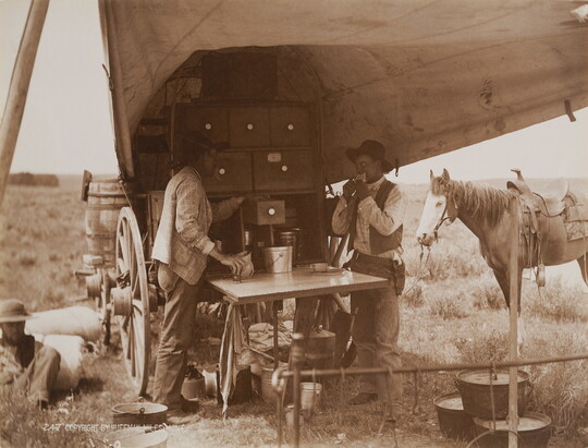 A sepia-toned photograph of two men eating and drinking from a chuckwagon as a horse stands in the background.