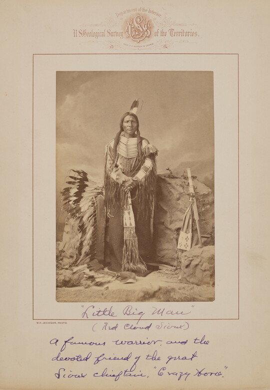A black-and-white formal portrait photograph of an Indigenous man in traditional clothing surrounded by a headdress and several Indigenous objects.