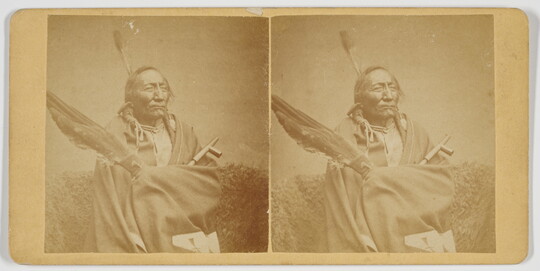A sepia-toned stereograph of an Indigenous man holding a group of large feathers and a pipe.