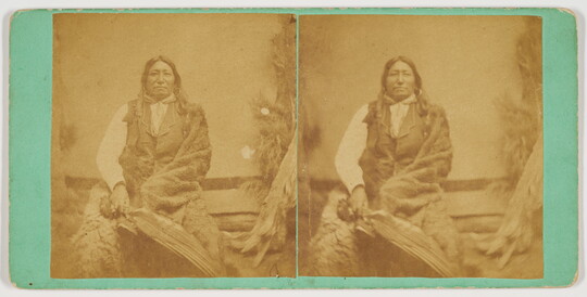 A sepia-toned stereograph of an Indigenous man wearing a vest and wrapped in a blanket, holding a feathers.