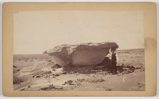 A sepia-toned photograph of a man standing next to a bowl-shaped rock formation in the desert.