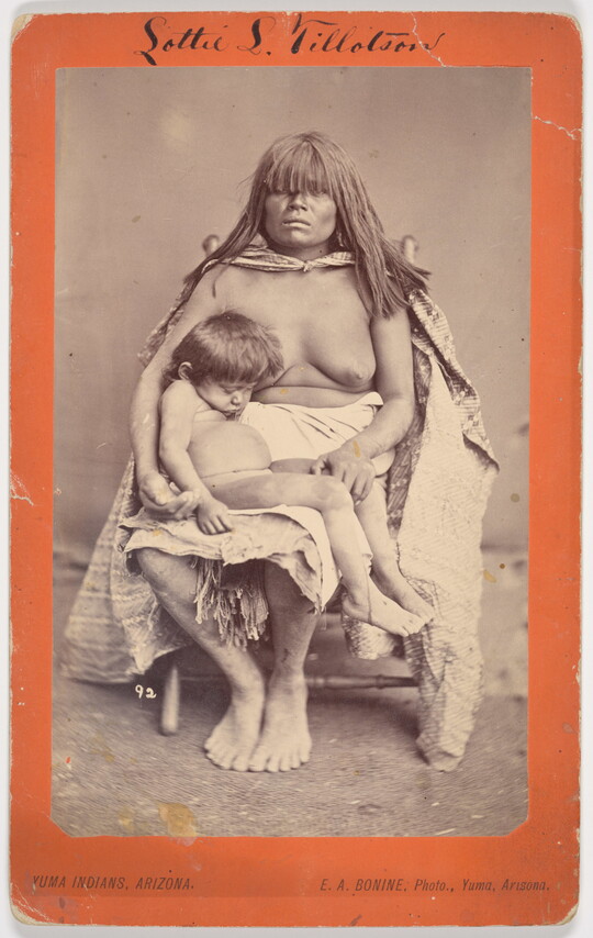 A black-and-white photograph of a seated Indigenous woman with long hair and bangs that almost cover her eyes, a blanket around her topless torso, and a sleeping child on her lap.