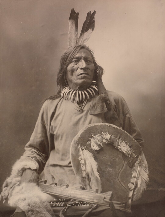 A black-and-white formal portrait photograph of a seated Indigenous man in traditional clothing holding a shield.