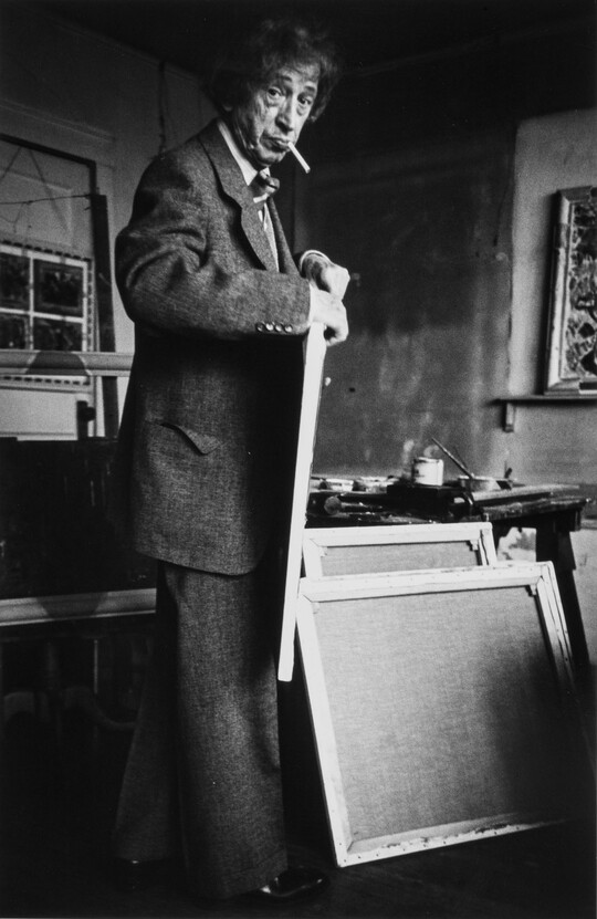 A black-and-white photograph of an older White man with a cigarette in his mouth holding a canvas in an artist's studio.