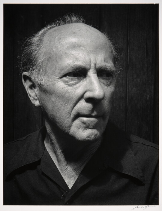 A black-and-white photograph of the head and shoulders of an older balding White man wearing a dark button-down shirt, head turned slightly.