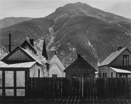 A black-and-white photograph of clapboard houses and a church at the base of a densely wooded mountain.