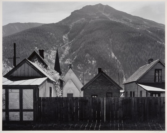 A black-and-white photograph of clapboard houses and a church behind a wood fence at the base of a densely wooded mountain.
