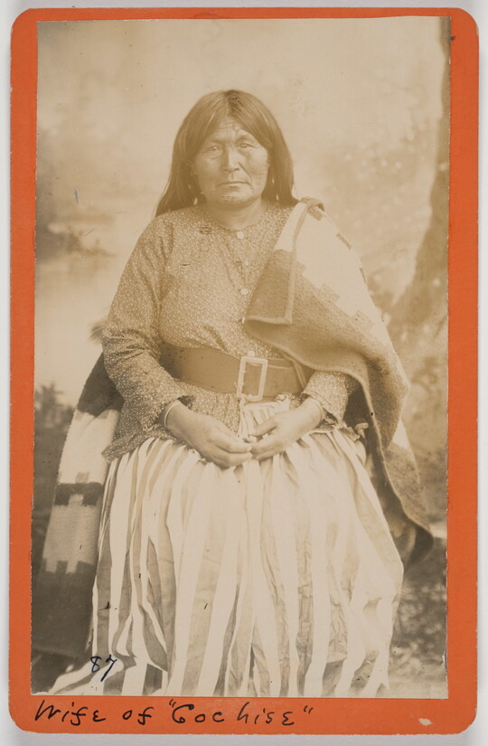 A sepia-toned formal portrait photograph of an Indigenous woman wearing a dress with a belt, and a blanket draped on her shoulders.