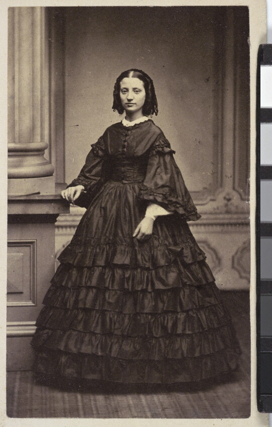 A black-and-white studio photograph of a White woman, dark hair styled in banana curls, wearing a long sleeved, full length, dark ruffled dress.