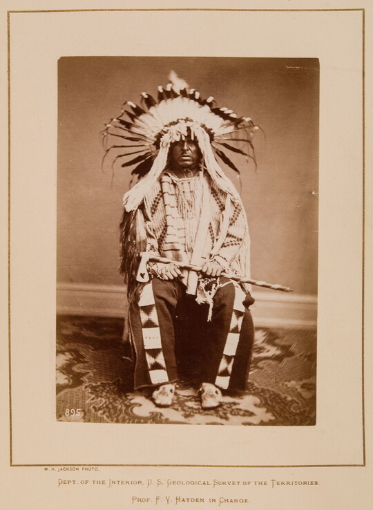 A black-and-white formal portrait photograph of a seated Indigenous man in full, traditional regalia with a headdress and tomahawk.