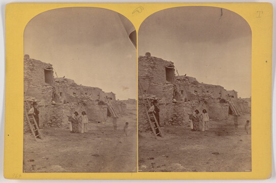 Two almost identical side-by-side black-and-white photographs of several Indigenous people at a multi-level Native American pueblo.