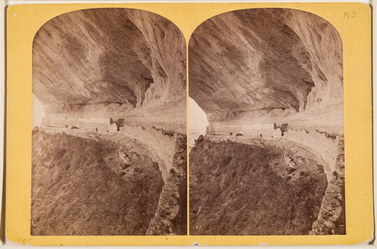 Two almost identical side-by-side sepia-toned photographs of a ruined stone structure built under the overhang of a cliff.