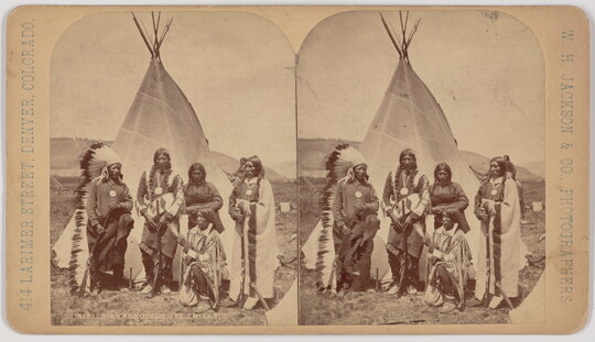 A black-and-white stereograph of five Indigenous people in traditional regalia standing in front of a tipi.