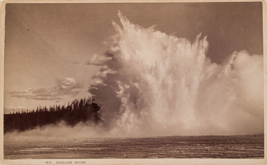 A sepia-toned photographic postcard of a large geyser erupting from muddy ground near evergreen trees.