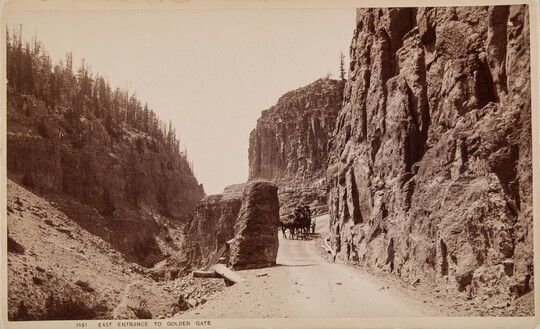 A sepia-toned photographic postcard of horses pulling a wagon on a dirt road between tall cliffs and rock formations.