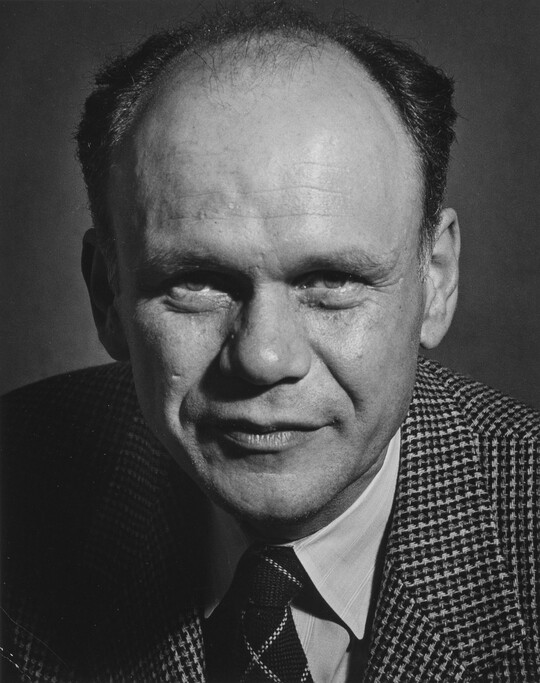 A black-and-white portrait photograph of a balding White man wearing a houndstooth blazer and striped tie.