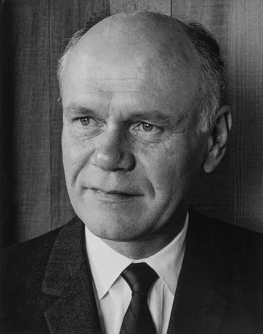 A black-and-white portrait photograph of a mostly bald White man gazing to the side wearing a dark jacket, dark tie, and white shirt.